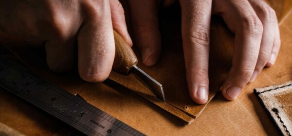 Close up of a shoemaker or artisan worker hands. Leather craft tools on old wood table. Leather craft workshop. ALVA artisan hand-cutting full-grain leather with a traditional leather knife in the Alva Leather Co. workshop in Lisbon, Portugal.